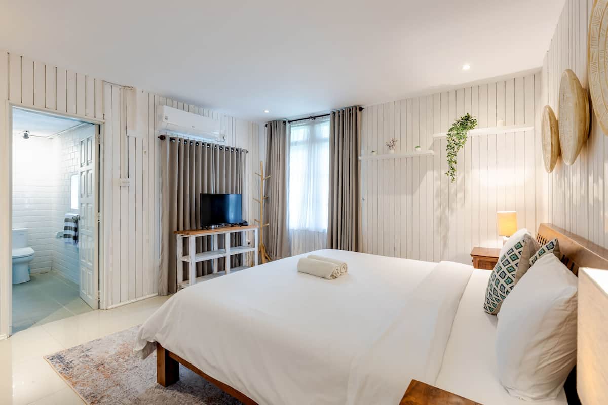 Quarto com cama grande, televisão em suporte, mesa de cabeceira com abajur e vista para o banheiro.