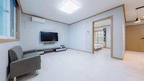 Una sala de estar moderna y bien iluminada con un televisor de pantalla plana, un sofá gris y una puerta de madera que da a otra habitación.