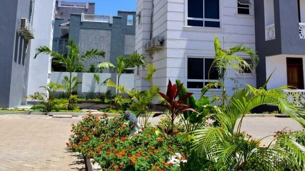 A modern residential building with multiple floors, balconies, and a paved pathway with flowering plants.