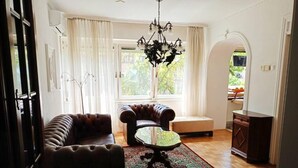 Ein Wohnzimmer mit einem braunen Ledersofa, einem Glastisch und einem gemusterten Teppich. Eine Kristallleuchter hängt von der Decke.