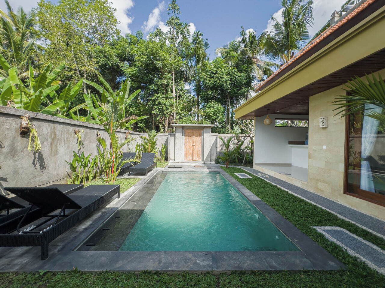 A rectangular swimming pool with a wooden door in the center, surrounded by lush greenery and a small garden area.