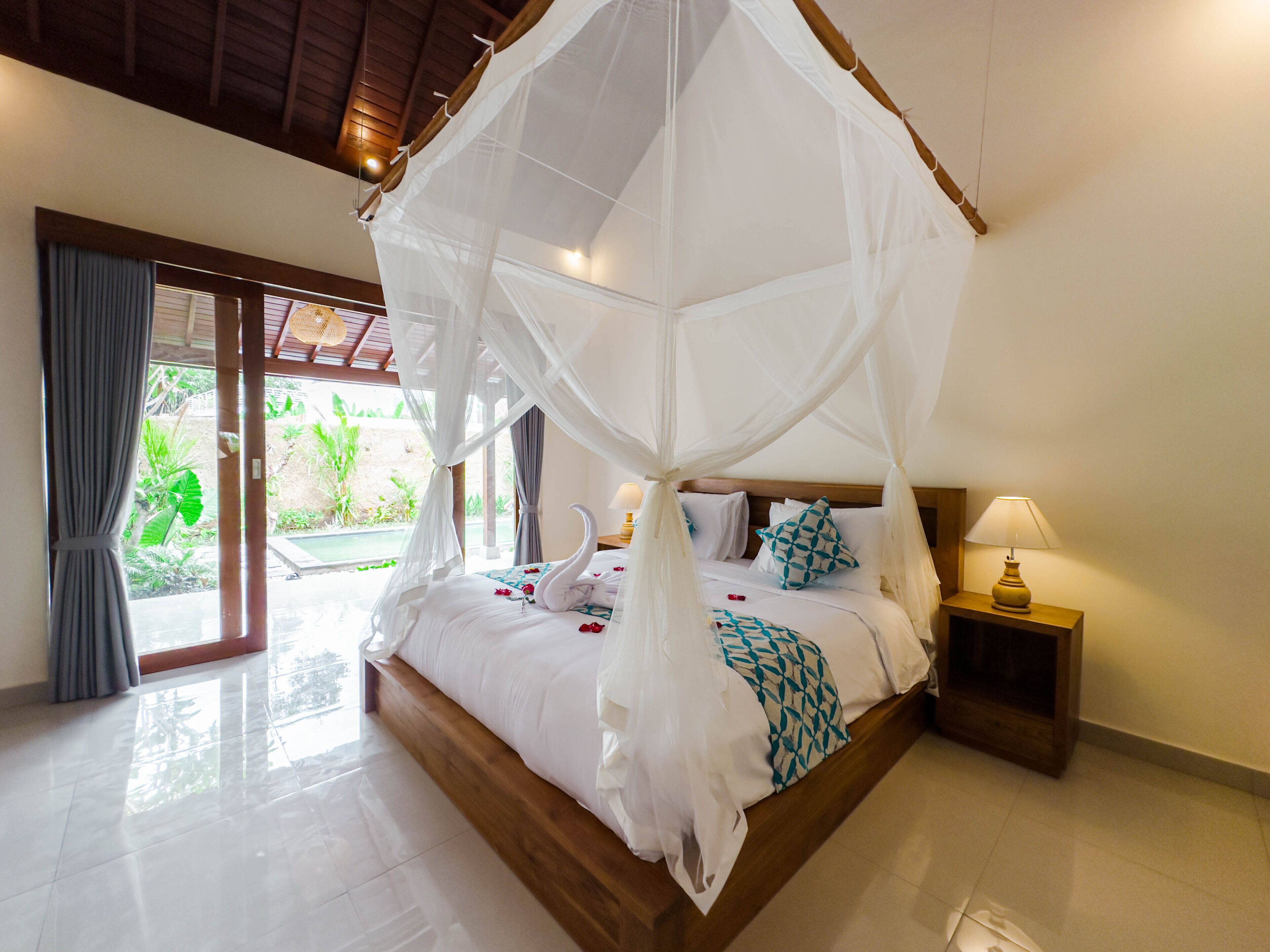 A spacious bedroom with a canopy bed, a wooden headboard, a bedside table with a lamp, and a view of the garden through the glass door.