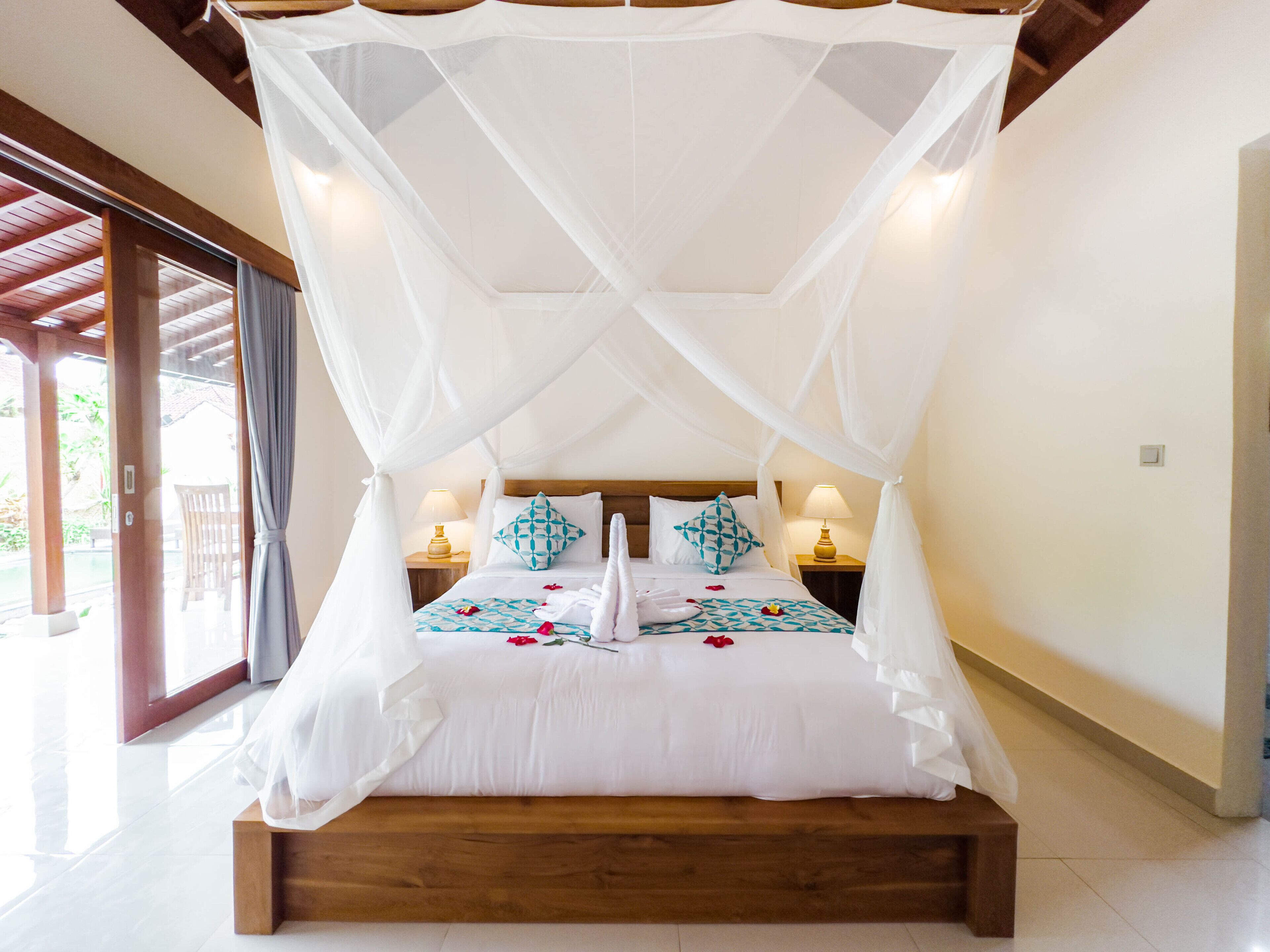 Una cama con dosel y cuatro columnas con sábanas blancas y almohadas decorativas, dos mesitas de noche con lámparas y vistas a un espacio exterior a través de una puerta de cristal.