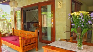 A wooden bed with a red mattress, a wooden table with a vase of flowers, and a glass door leading to a room. - Tutik Private House by Supala (Ubud)