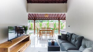 A modern living room with a TV, wooden furniture, a grey sofa, and a view of a garden and pool. - Tutik Private House by Supala (Ubud)