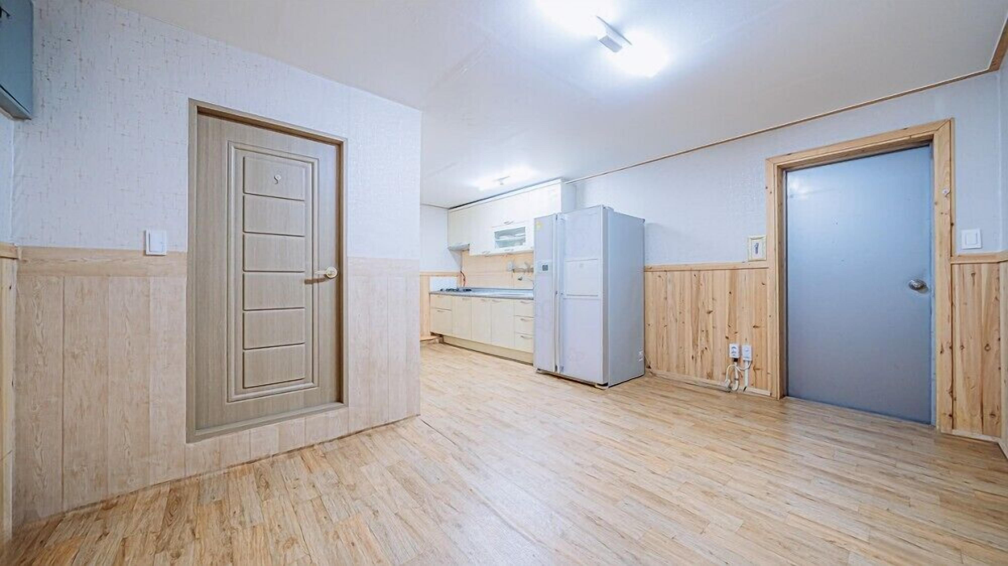A spacious room with wooden flooring, a built-in kitchen with a refrigerator, and a door with a panel design.