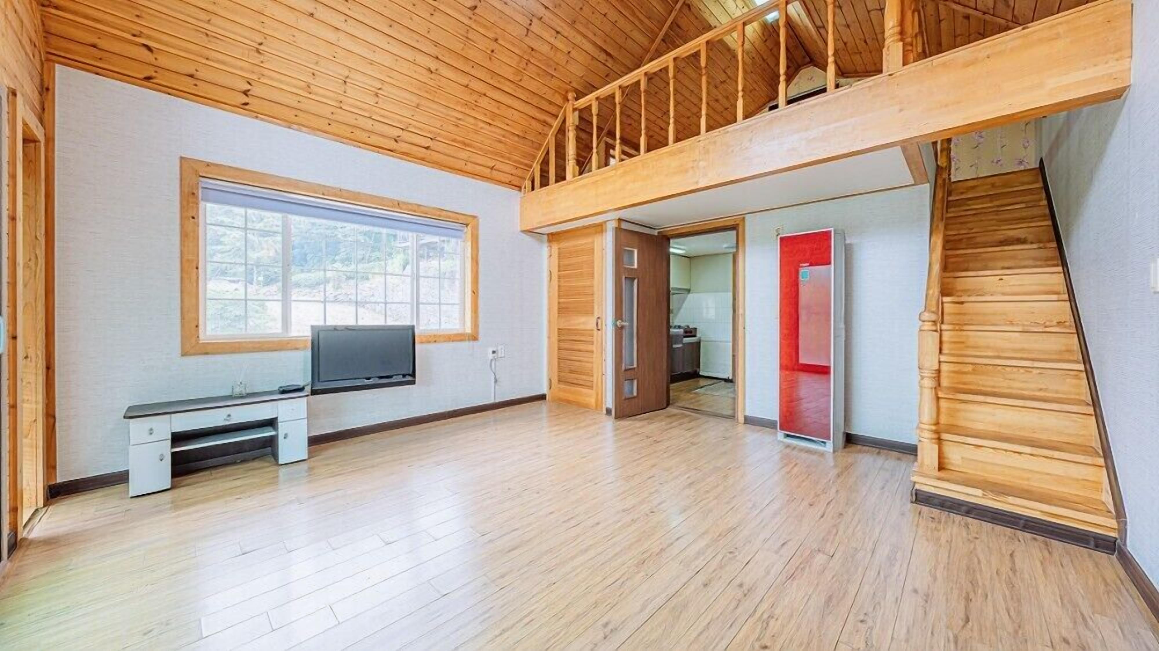 A spacious room with a wooden floor, a TV on a stand, a balcony, and a red door leading to another room.