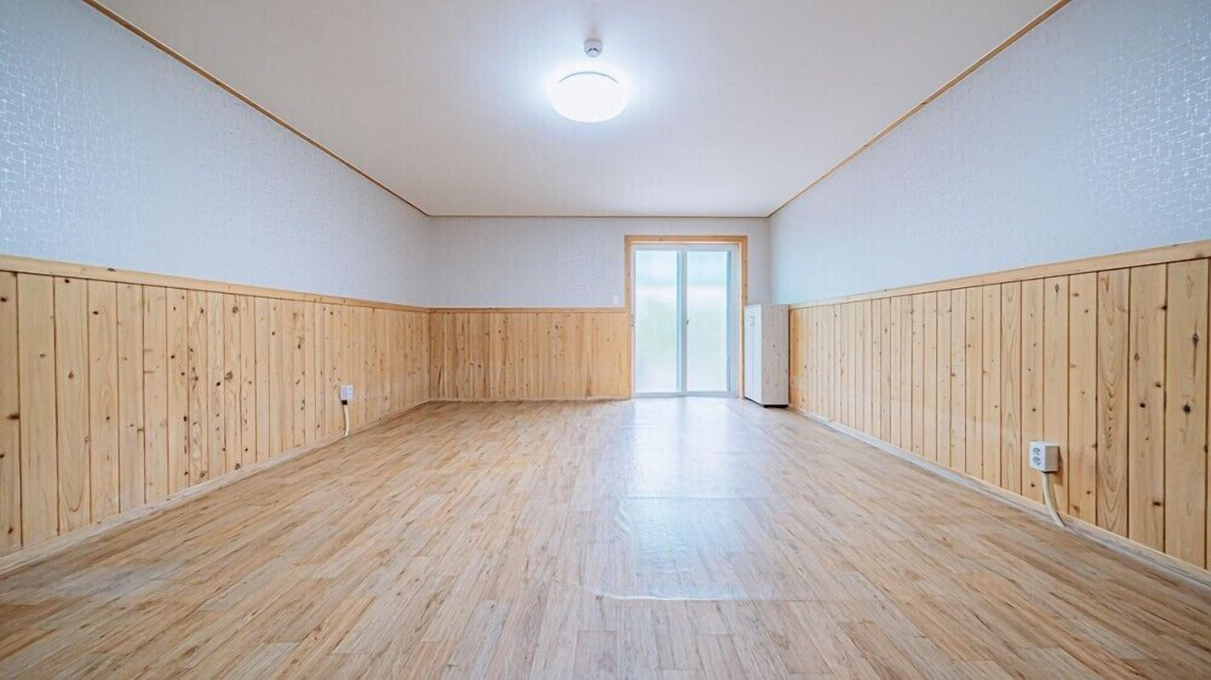 En rummelig stue med træpaneler på vægge og gulv, en loftlampe og en dør der fører til et andet værelse.