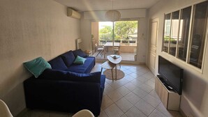 A living room with a dark blue sofa, a small round table, and a TV on a stand. - Très Beau T2 Neuf, Calme, Terrasse, au Cœur de Sanary + box Fermé en Sous-sol (Sanary-sur-Mer)