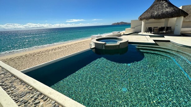 Una piscina blava i cristal·lina amb una banyera d'hidromassatge, una platja i una construcció amb teulada de palla.