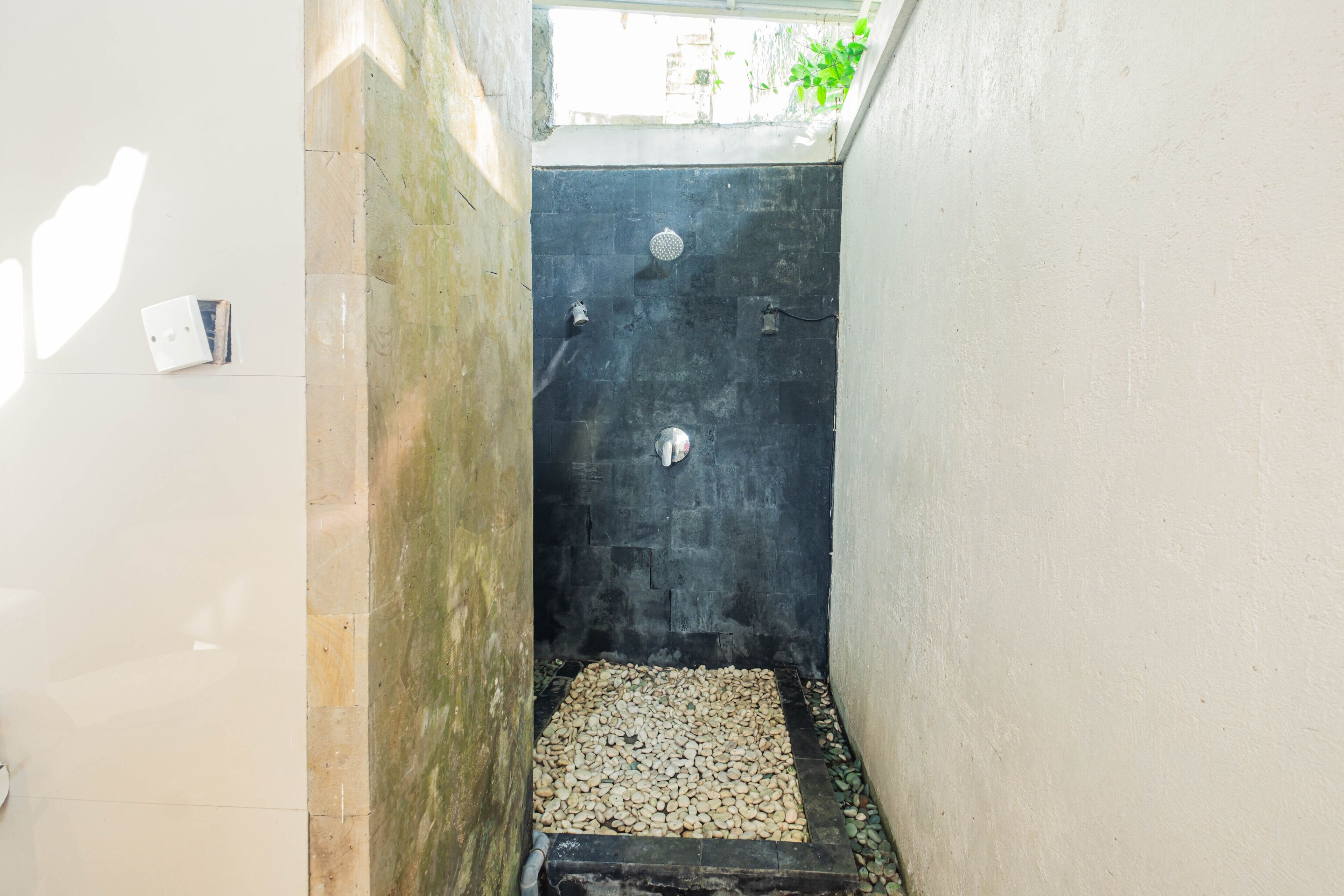 Un espacio de ducha pequeño y cerrado, con una pared de azulejos oscuros, piso de azulejos blancos y una pequeña ventana en la parte superior.