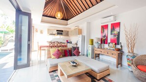 A modern living room with a glass door leading to a pool, a sofa with colorful cushions, a wooden coffee table, and a dining area with a table and chairs. - Kakul Villa F by Supala (Ubud)