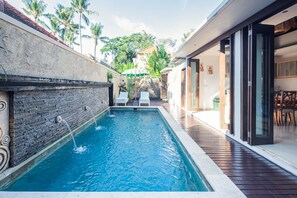 A swimming pool with two lounge chairs, a stone wall, and a wooden deck. - Kakul Villa F by Supala (Ubud)
