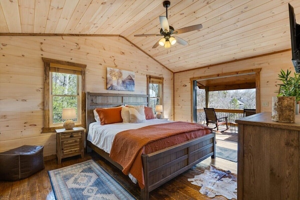 Un dormitorio de cabaña de madera con una cama, un ventilador de techo, una ventana, una mesita de noche y una alfombra.