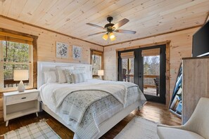 A wooden cabin bedroom with a bed, nightstands, a ceiling fan, and a view through a door to an outdoor deck.