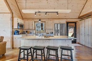 Interior de una cabaña de madera con una isla en la cocina, taburetes de bar y un ventanal grande.