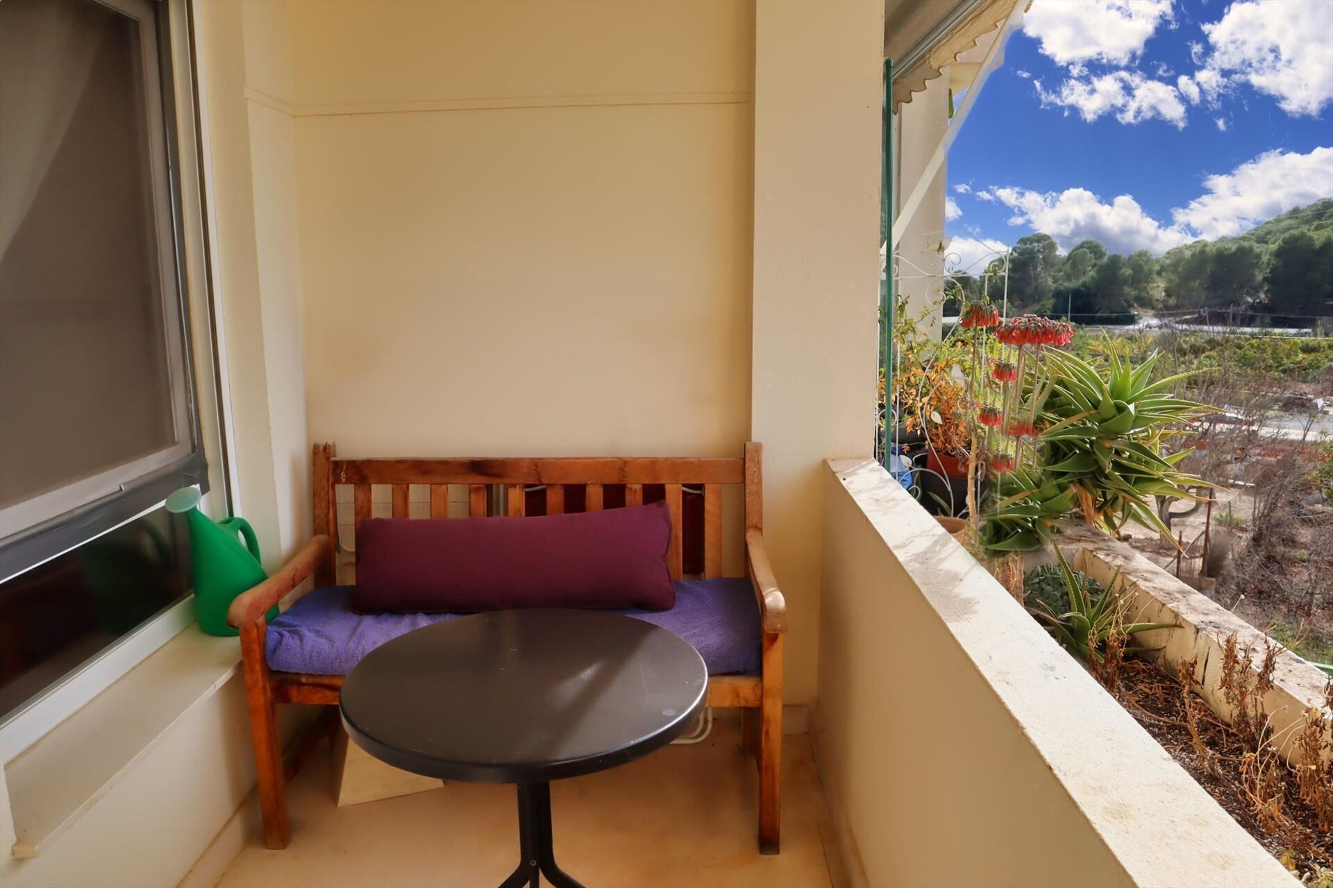 Un balcon avec un banc en bois, une petite table ronde et une vue sur un jardin planté d’arbustes et d’arbres.