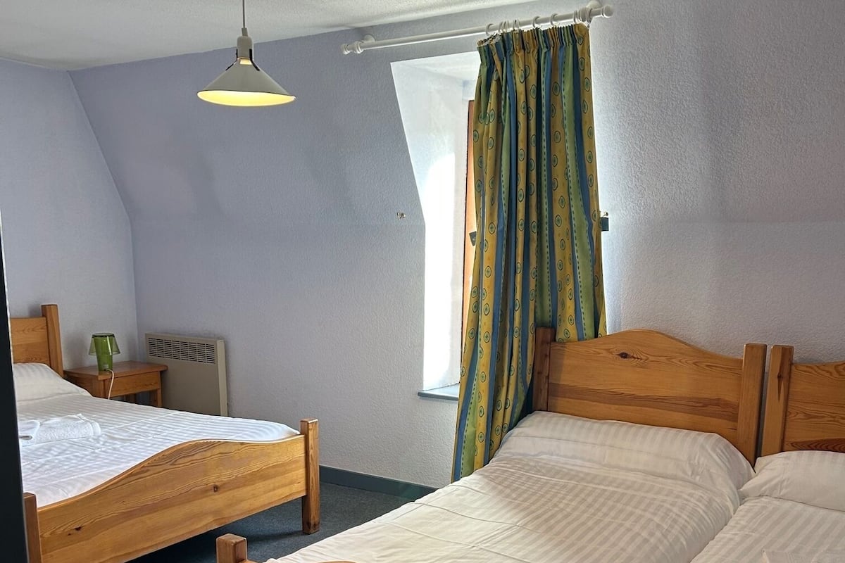 A room with two wooden beds, a small table with a lamp, a window with curtains, and a ceiling light.