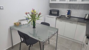 Een moderne keuken met witte kastjes, een eettafel met glazen blad, een zwarte stoel, een broodrooster, een waterkoker en een vaas met bloemen.