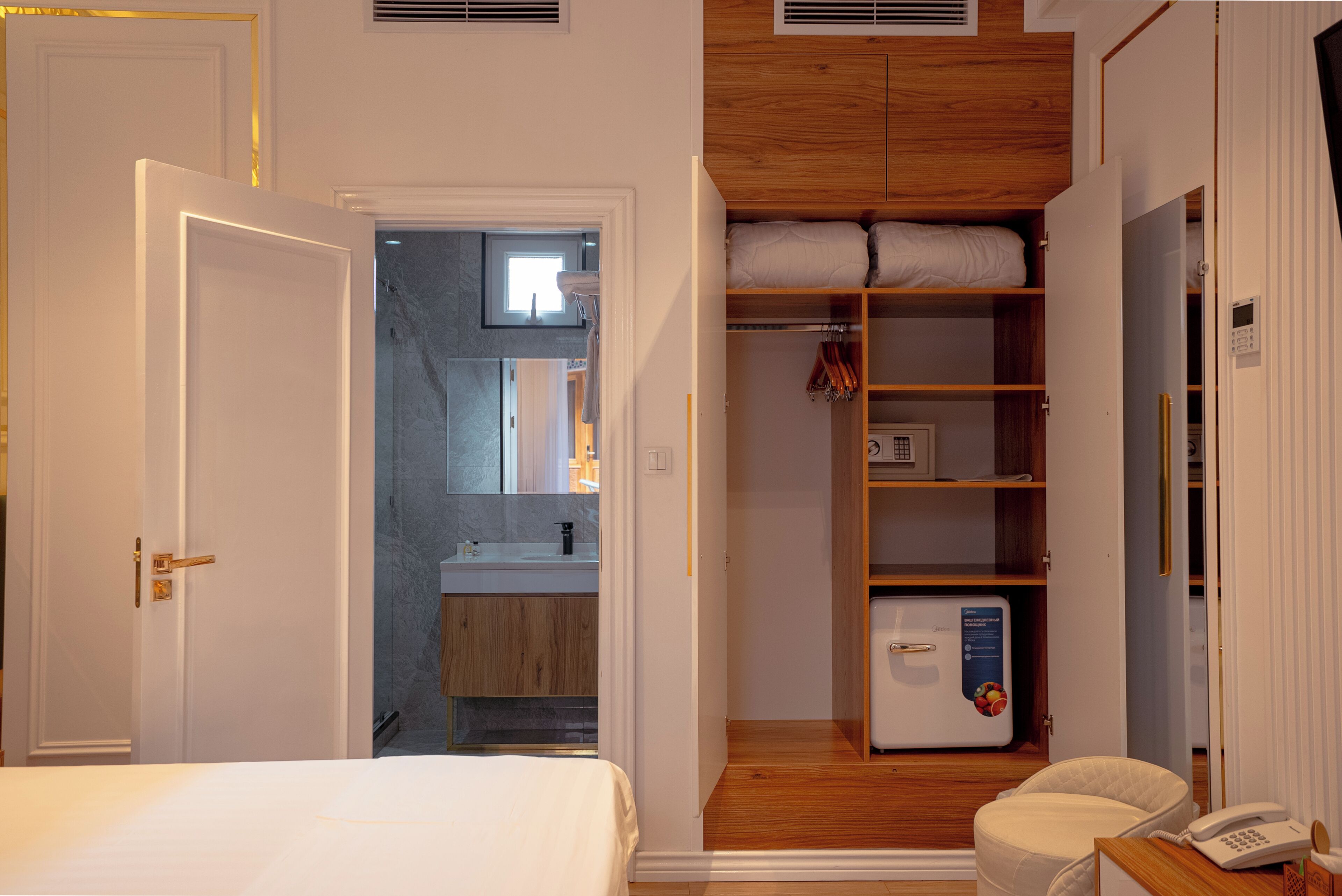 A hotel room with a bed, a wooden wardrobe, a small bathroom with a mirror and sink, and a white telephone on a wooden table.