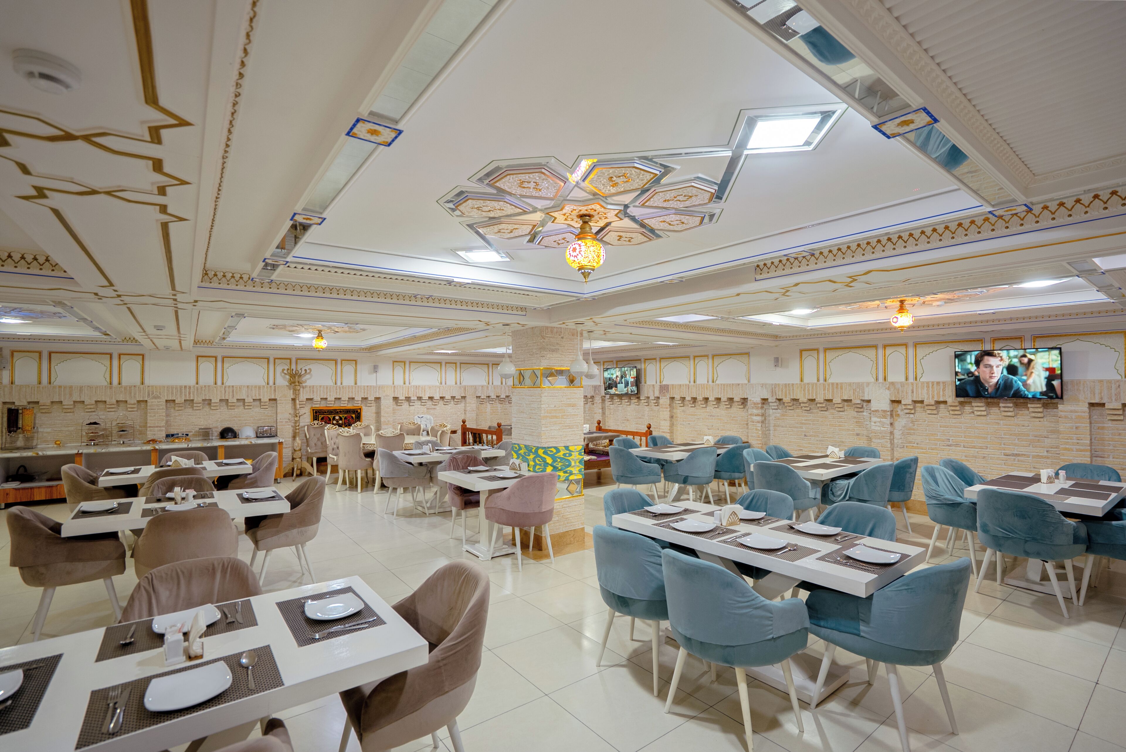 A spacious restaurant with multiple dining tables and chairs, a decorative ceiling, and a television screen.
