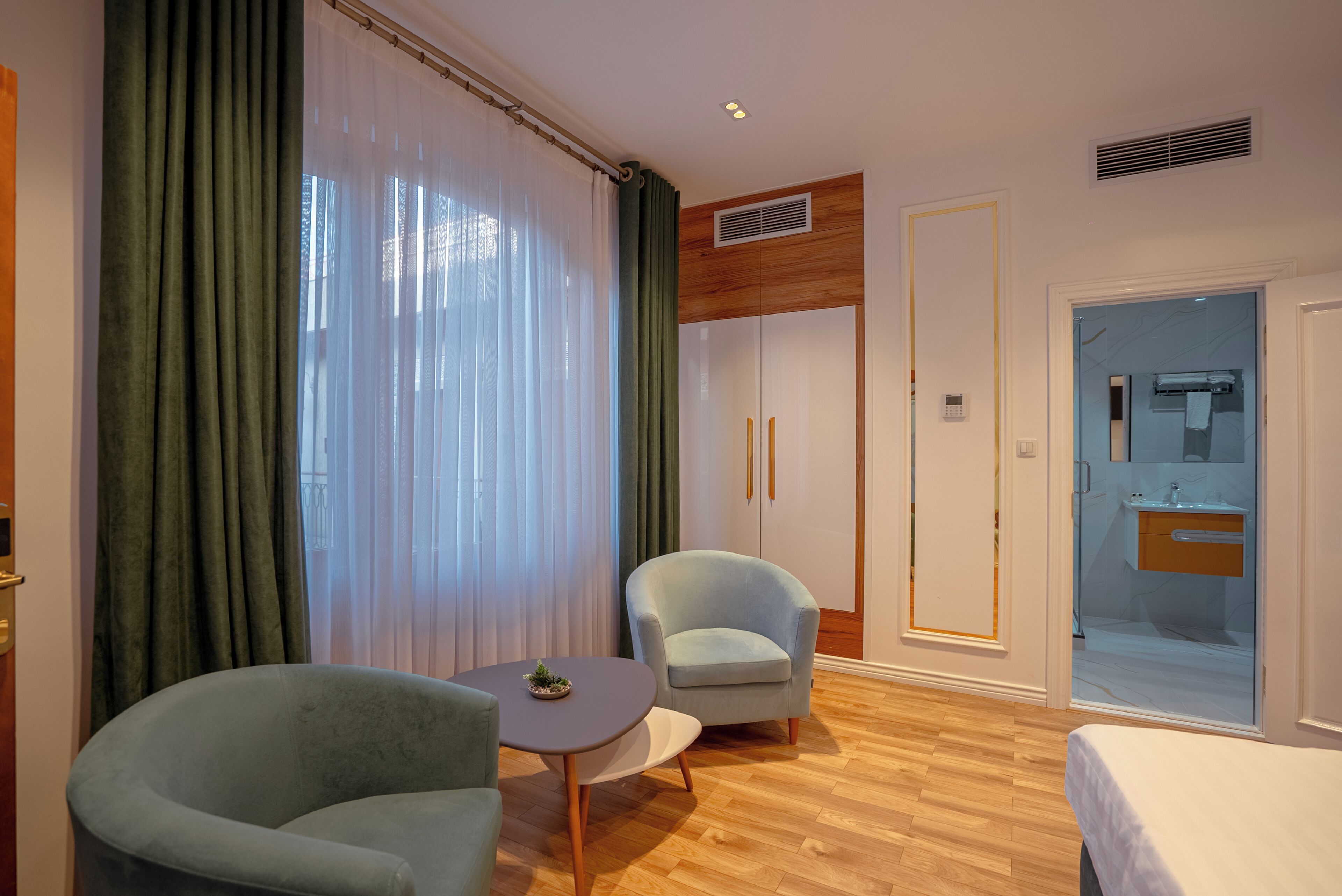 A hotel room with a wooden floor, a bed, a small round table with a plant, two armchairs, a large window with sheer curtains, and a bathroom visible through a glass door.