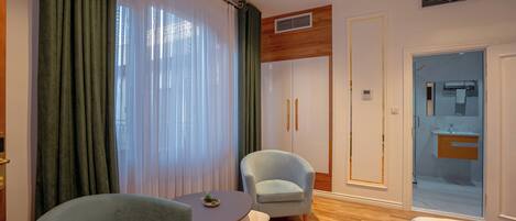 A hotel room with a wooden floor, a bed, a small round table with a plant, two armchairs, a large window with sheer curtains, and a bathroom visible through a glass door.