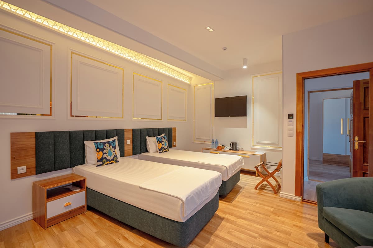 Quarto de hotel com duas camas, uma televisão, uma mesa de cabeceira de madeira, uma cadeira e uma porta que dá para outro quarto.