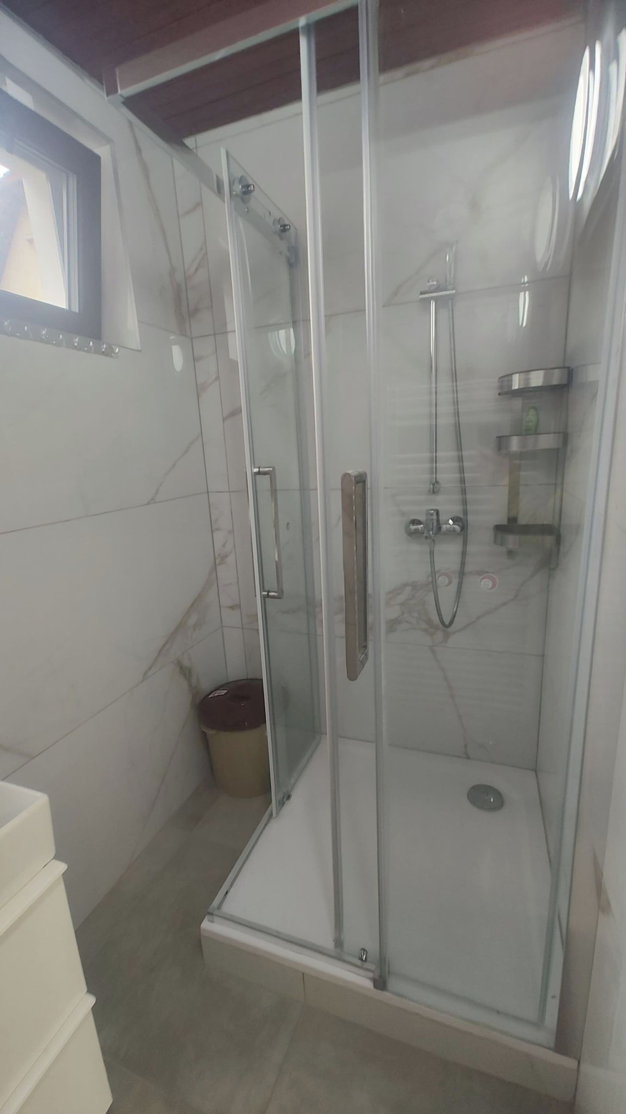 Eine Duschkabine mit Glaswänden, einem Griff, einem kleinen Mülleimer und einem Fenster darüber.