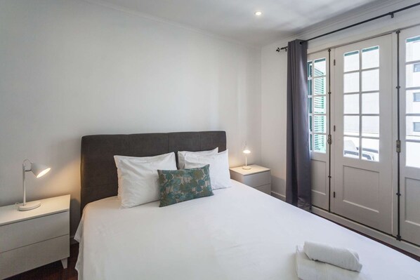 A neatly made bed with white linens and a grey headboard, two bedside lamps, and a window with curtains.