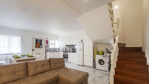 A modern living room with a brown sectional sofa, a dining area with a table set for four, a kitchen with white cabinets and a washing machine, and a wooden staircase. - Atlantic City - Marina (Ponta Delgada)