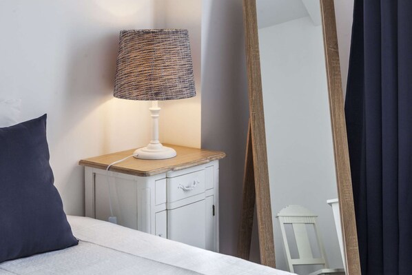Een bed met een wit hoofdeinde, een nachtkastje met een lamp, een donkerblauwe kussens en een spiegel die een kamer weerkaatst met een houten deur en een witte kast.