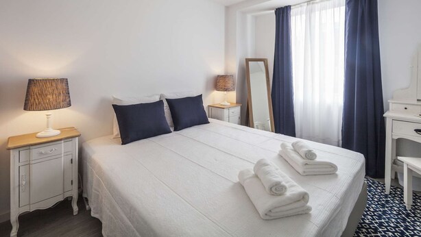 A hotel room with a neatly made bed, two bedside lamps, a large mirror, a small desk with a chair, and a window with curtains.