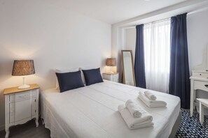 A hotel room with a neatly made bed, two bedside lamps, a large mirror, a small desk with a chair, and a window with curtains.
