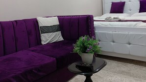 A modern bedroom with a purple sofa, a white bed with purple pillows, a small table with a plant, and wall decorations including a clock and a mirror. - Lux Suites Mattlet Studio Apartments TRM (Nairobi)
