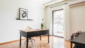 Quarto moderno e bem iluminado, com piso de madeira, uma mesa grande e uma cadeira. Há um vaso de planta sobre a mesa e um quadro na parede.