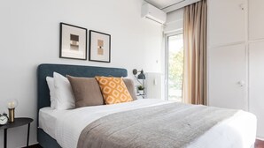 Une chambre à coucher bien rangée, avec un lit, une table de chevet dotée d’une lampe et une fenêtre avec des rideaux.