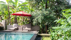A swimming pool with lounge chairs and a red umbrella, surrounded by lush greenery.