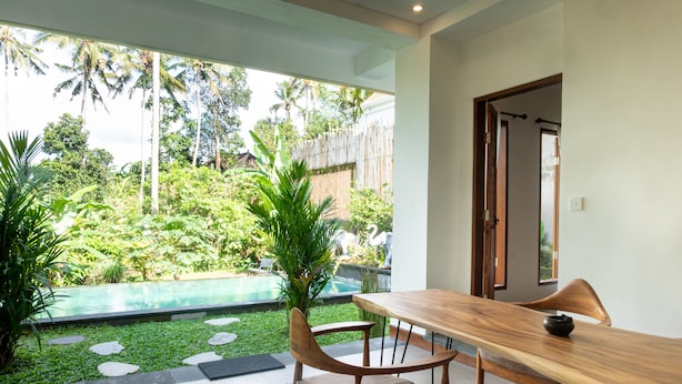 A dining area with a wooden table and chairs, a view of a swimming pool, and tropical vegetation.