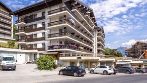 A multi-story hotel with balconies, a parking lot, and a mountain in the background. - Valaisia 55B - 5 pers - plein centre (Haute-Nendaz)