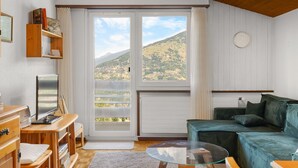 A room with a wooden ceiling, a green sofa, a glass table, a wooden TV stand, and a view of a hilly landscape through large windows. - Valaisia 55B - 5 pers - plein centre (Haute-Nendaz)