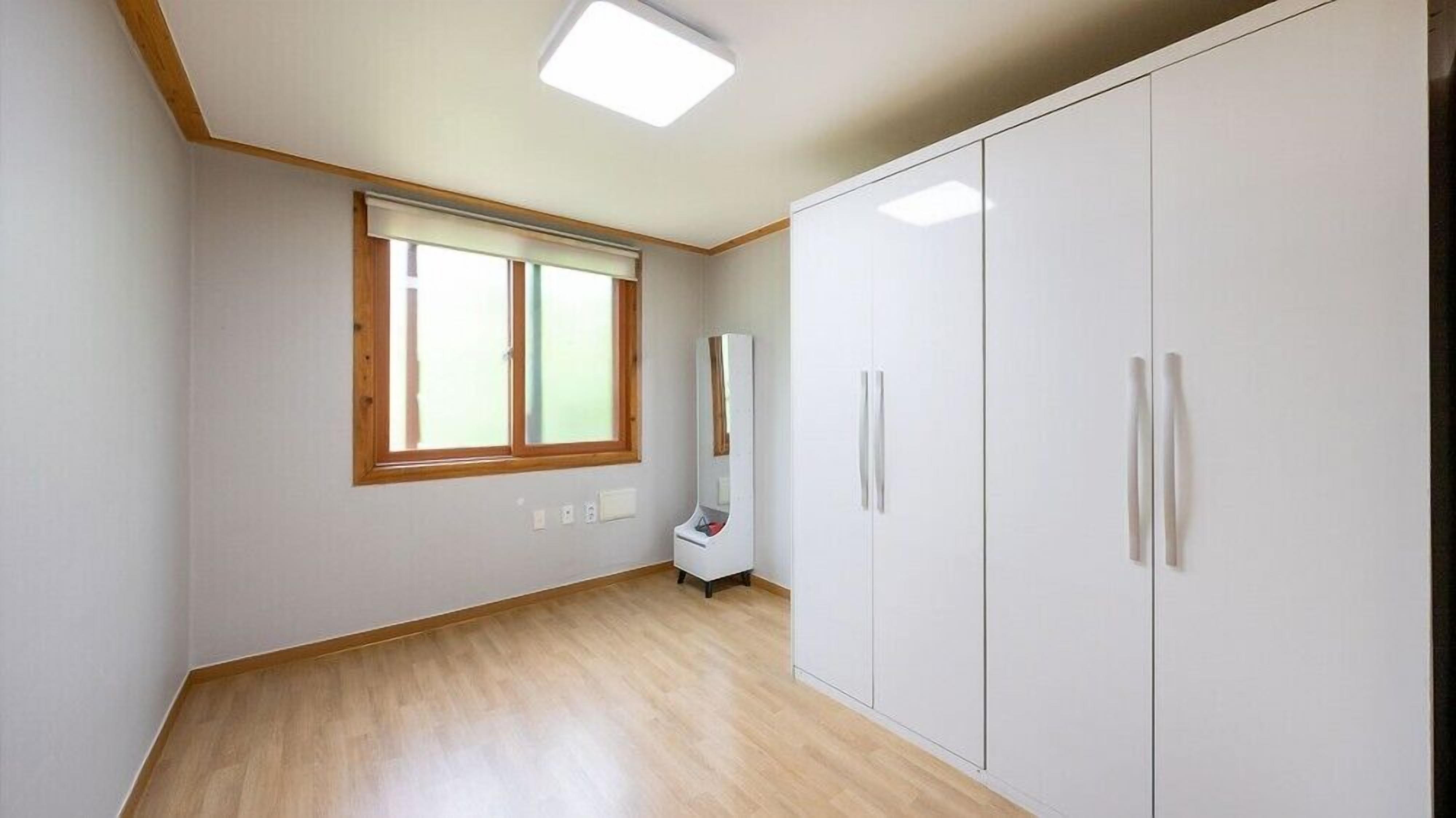 A room with a wooden floor, white walls, a large window with blinds, and a white wardrobe with two doors.