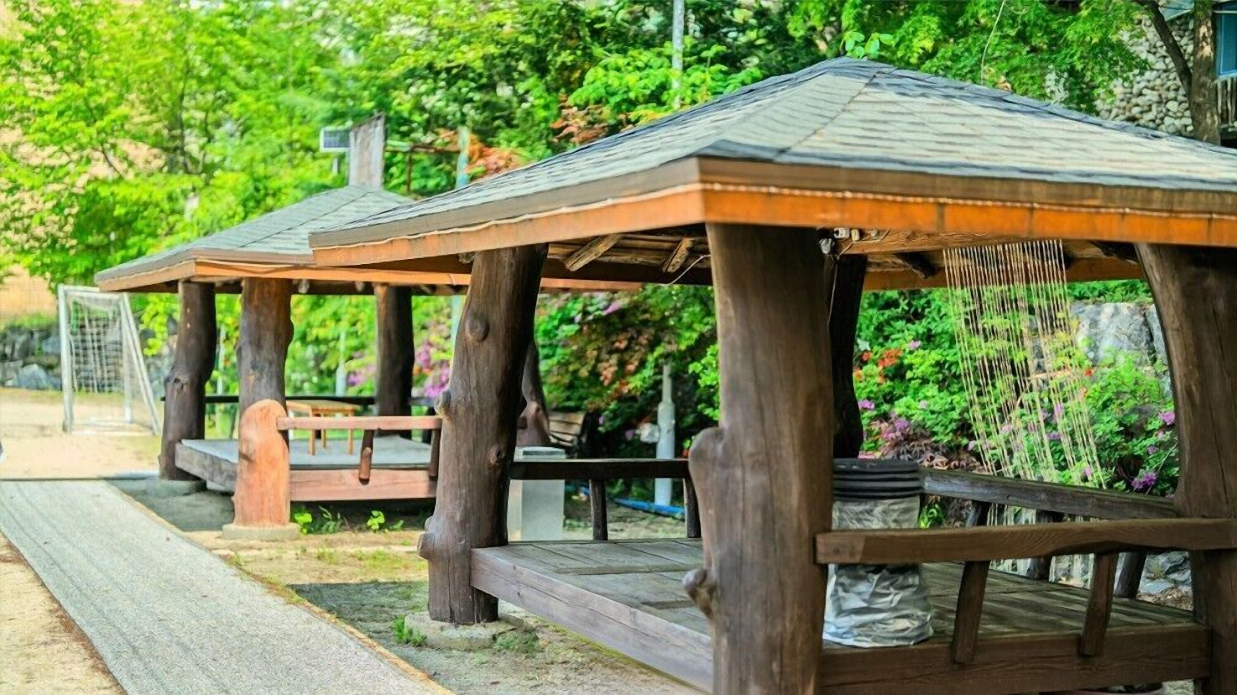 Deux pergolas en bois, dotées de toits à lattes, entourées de verdure et bordées d’un chemin pavé.