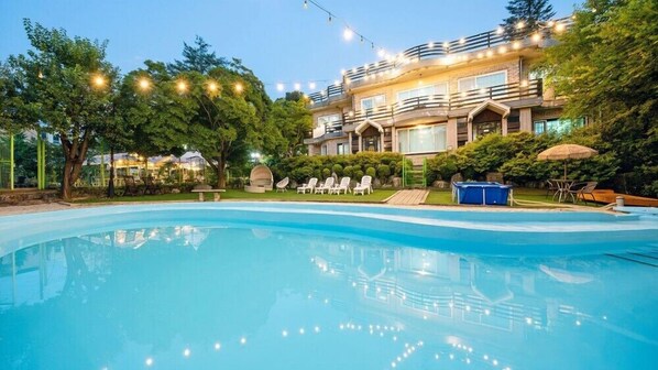 Una piscina con sillas de descanso y un jacuzzi, rodeado de árboles y un edificio con balcones.