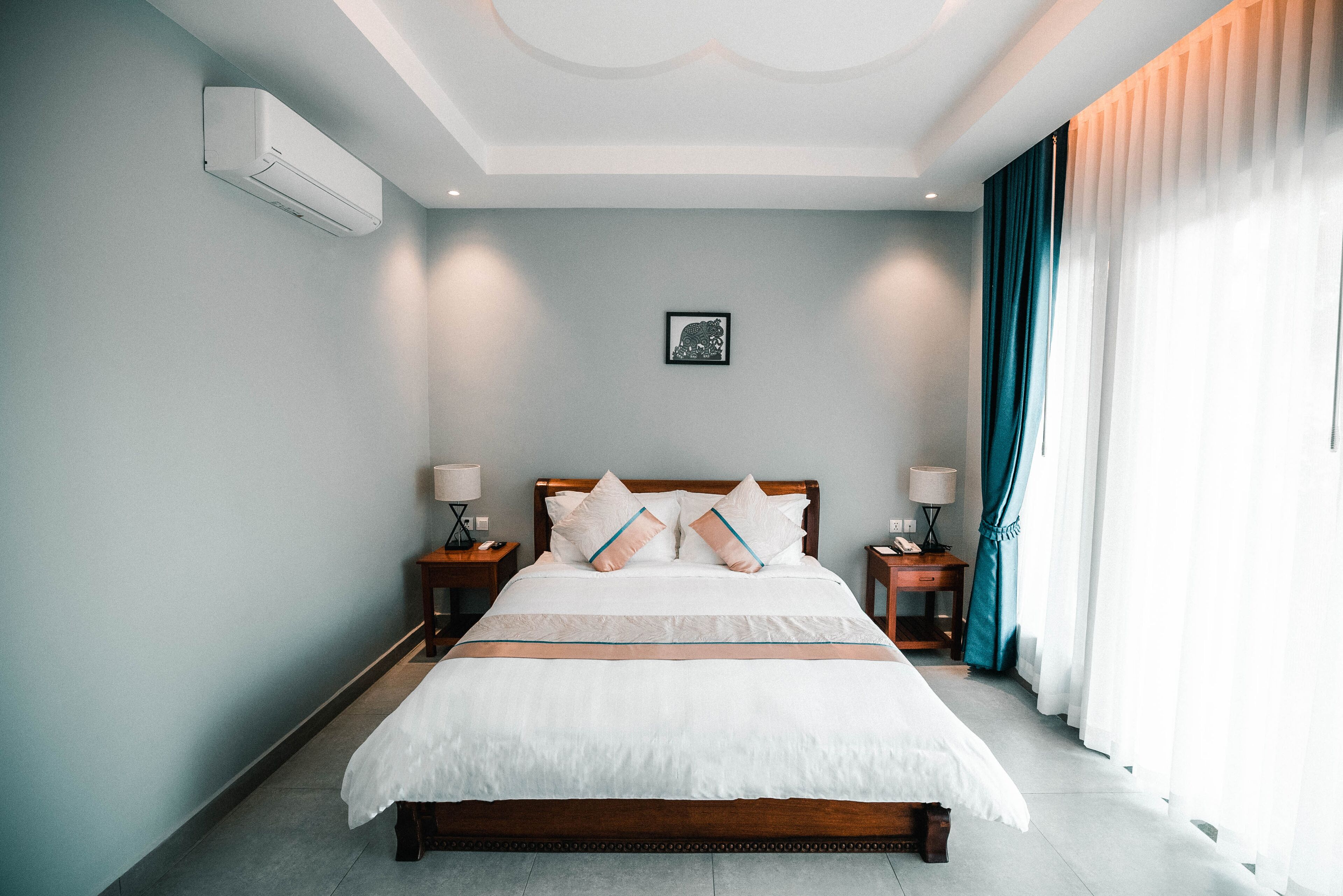 A neatly made bed with a wooden headboard, two bedside tables with lamps, a framed picture on the wall, and a window with curtains.