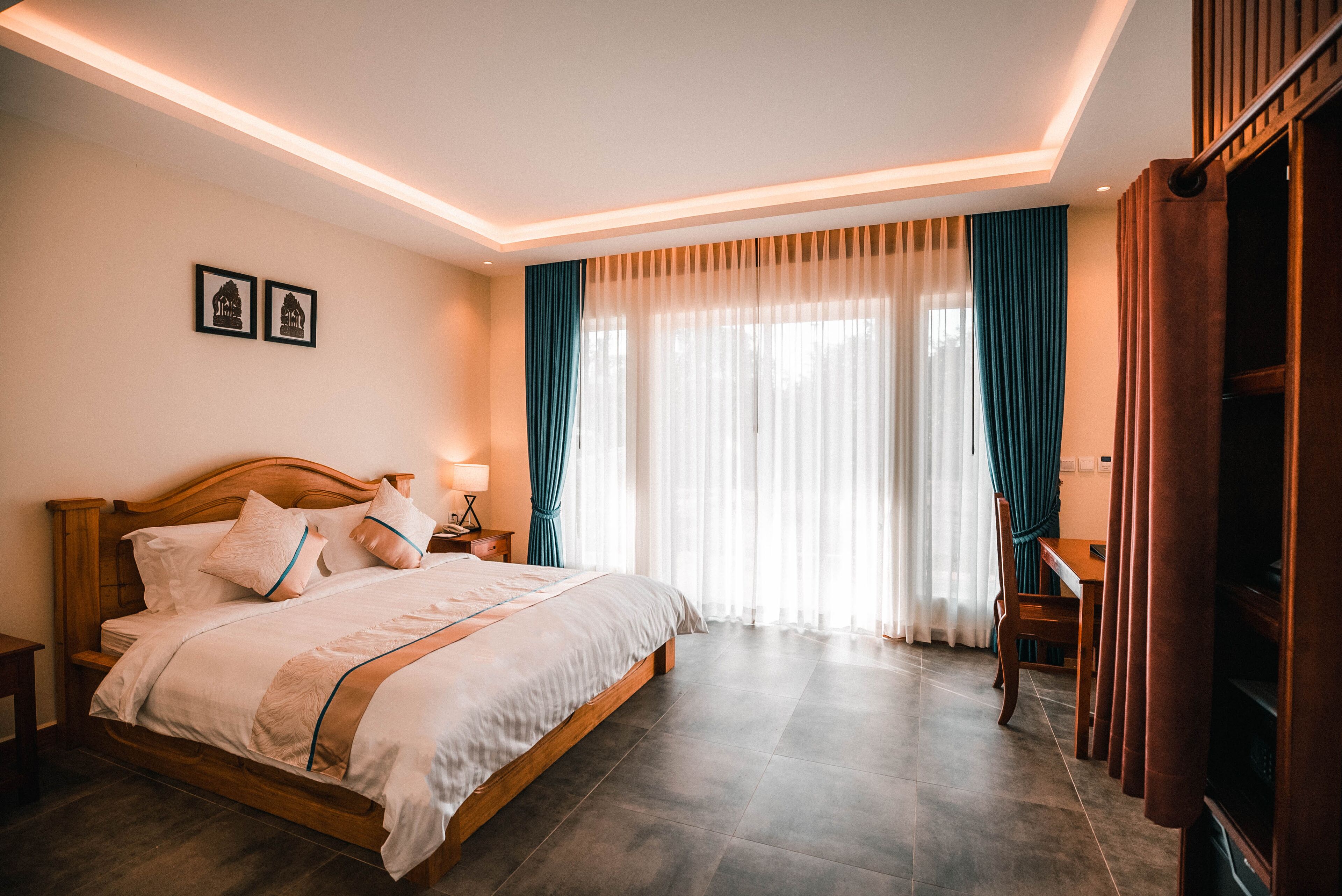 A neatly arranged bedroom with a large bed, a bedside table with a lamp, and a view of the outdoors through sheer curtains.