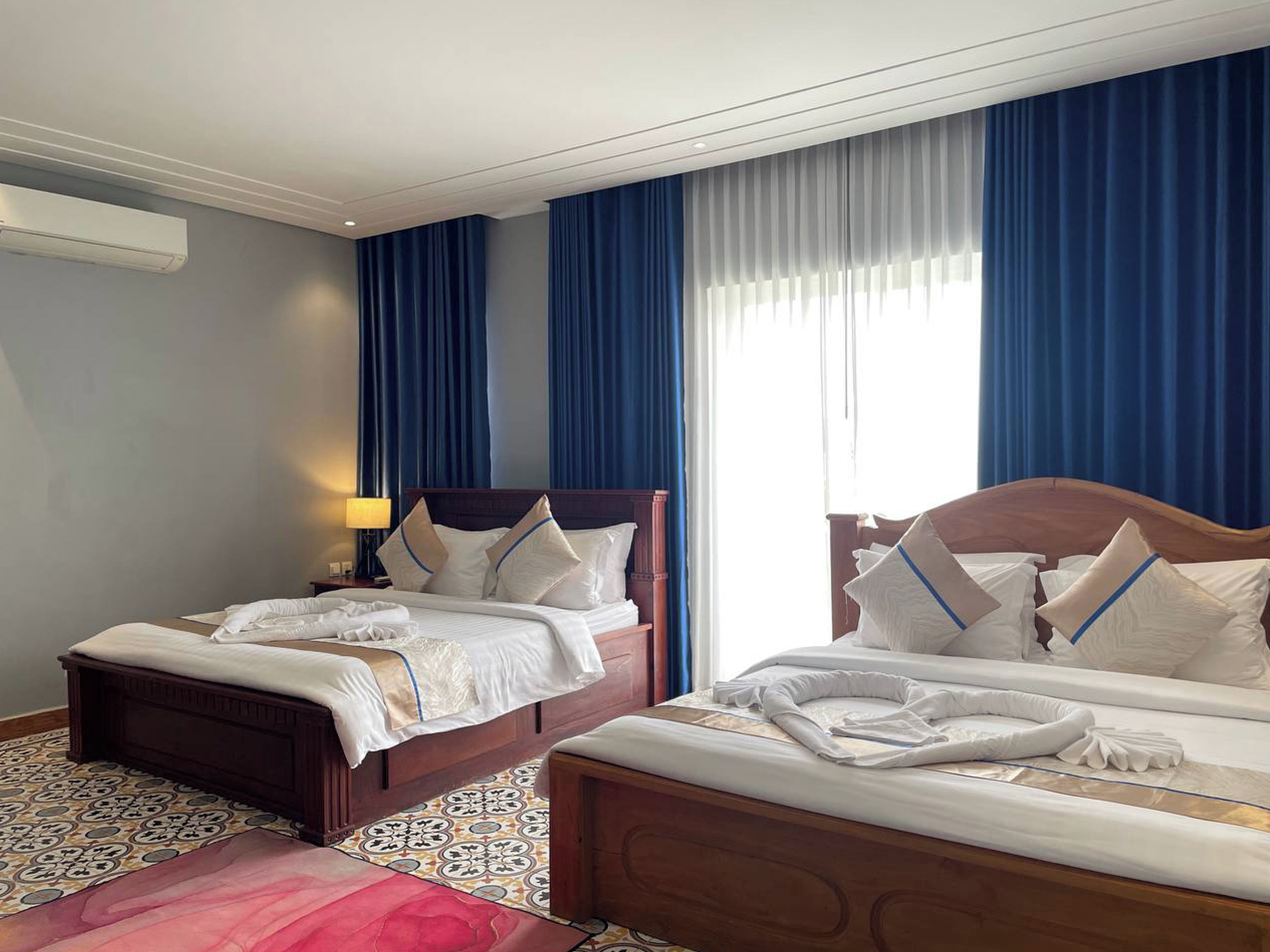 A hotel room with two beds, a patterned floor, and a window with blue curtains.