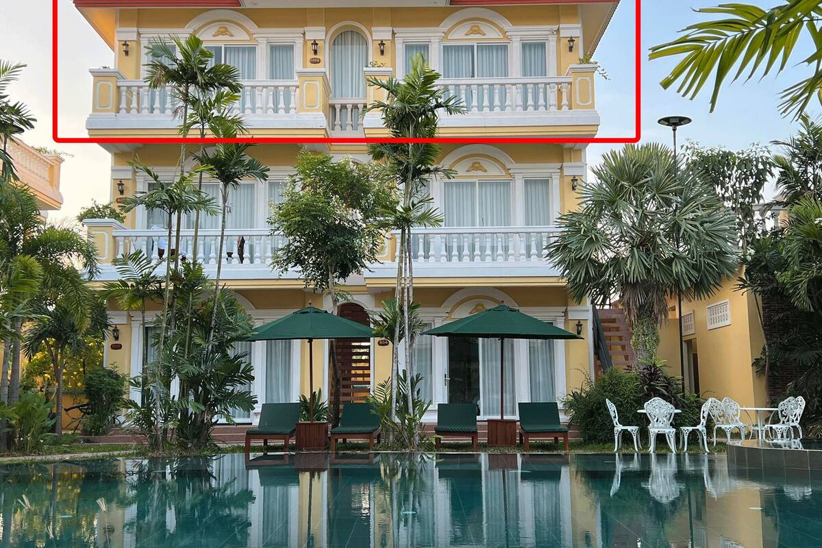 A two-story building with a balcony, surrounded by a pool and lounge area with chairs and umbrellas.