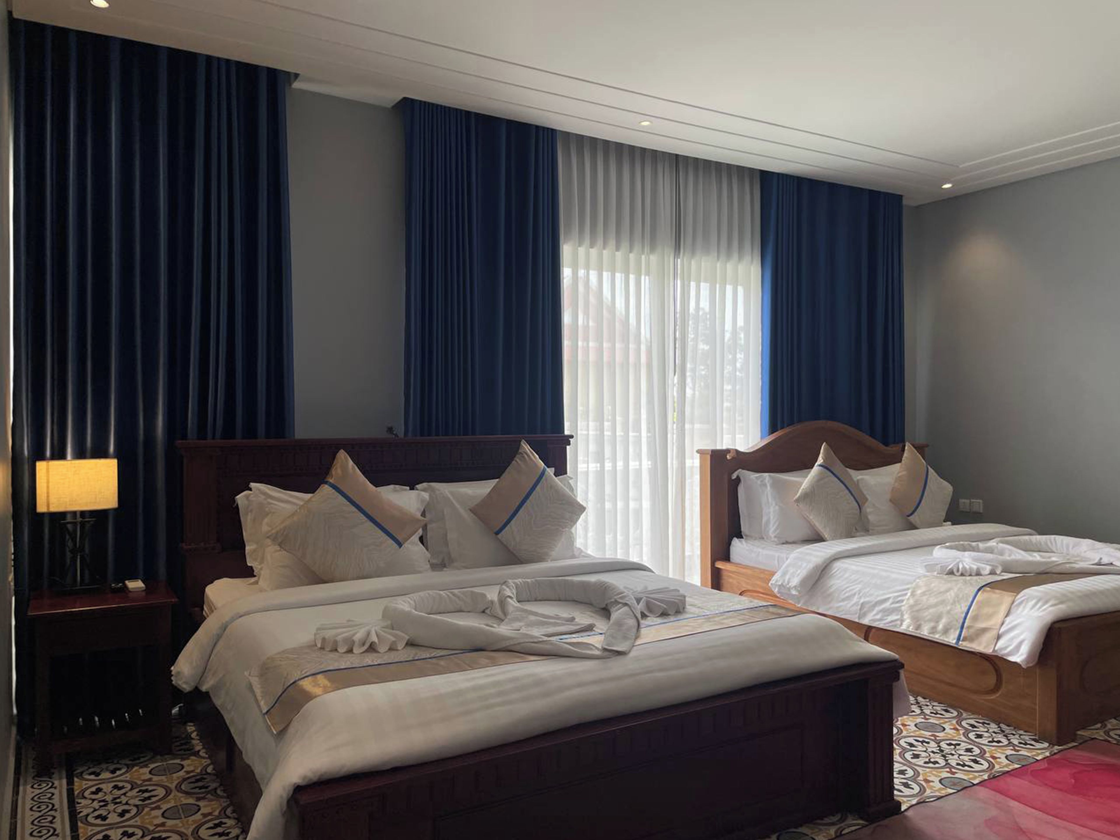 A hotel room with two beds, a nightstand, a lamp, and a large window with curtains.