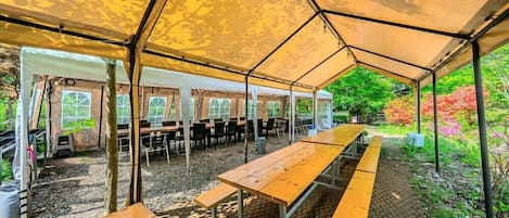 A covered outdoor seating area with tables and chairs, surrounded by a garden with trees and shrubs.